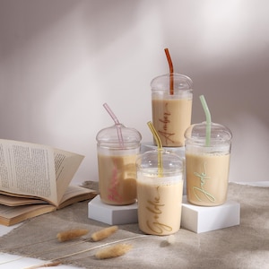 Pu&ograve; includere: Quattro bicchieri trasparenti con coperchi a cupola e cannucce in vetro colorato, riempiti con un liquido beige. I bicchieri presentano nomi in corsivo in colori coordinati: Amber, Goldie, Jade e un nome rosa sul bicchiere a sinistra.