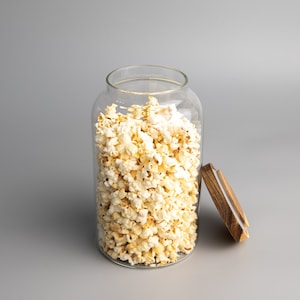 May include: A clear glass jar filled with popcorn, with a wooden lid sitting next to it on a grey background.