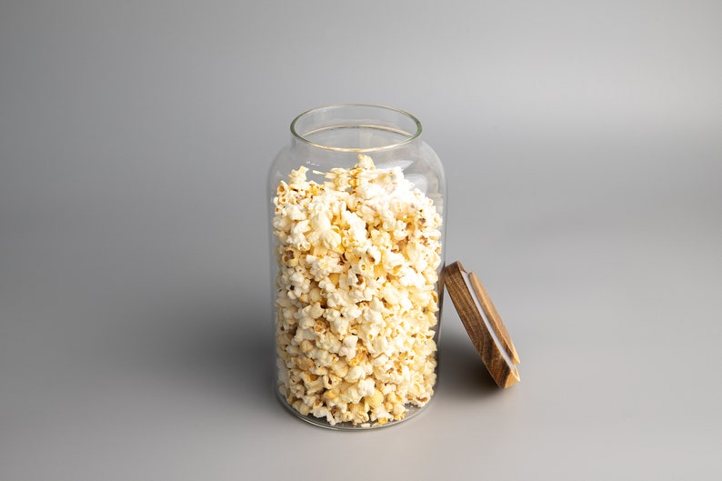 May include: A clear glass jar filled with popcorn, with a wooden lid sitting next to it on a grey background.