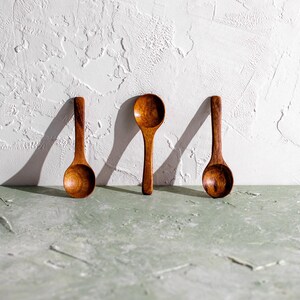 Pu&ograve; includere: Tre cucchiai di legno con ciotole arrotondate e manici lunghi sono disposti contro un muro bianco strutturato. I cucchiai sono fatti di legno dai toni caldi e l'illuminazione crea ombre. I cucchiai sono su una superficie verde chiaro.