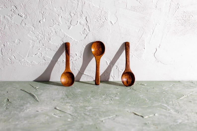 Pu&ograve; includere: Tre cucchiai di legno con ciotole arrotondate e manici lunghi sono disposti contro un muro bianco strutturato. I cucchiai sono fatti di legno dai toni caldi e l'illuminazione crea ombre. I cucchiai sono su una superficie verde chiaro.