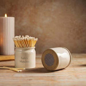 May include: A cream ceramic match holder and striker are displayed with wooden matches. The holder is labelled "Safety Matches". Lit candles and a ribbed candle are in the background, creating a warm, inviting atmosphere.
