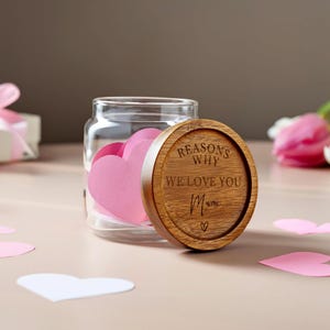 Könnte beinhalten: Ein klares Glasgefäß gefüllt mit rosa herzförmigen Notizen, mit einem Holzdeckel mit der Aufschrift "REASONS WHY WE LOVE YOU Mum". Das Glas steht auf einer hellrosa Oberfläche, mit Herz-Ausschnitten.