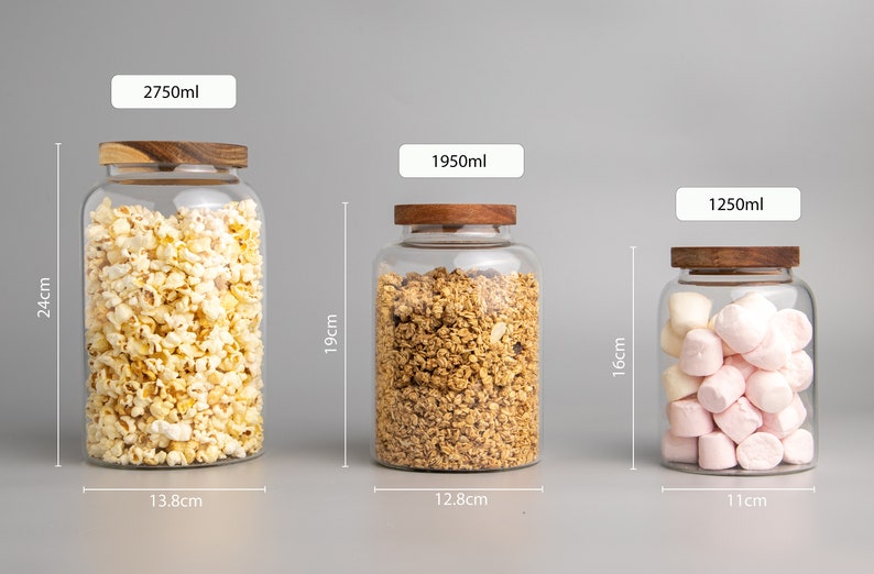 May include: Three glass storage jars with wooden lids, filled with popcorn, granola, and marshmallows. The jars are labelled with their capacity in millilitres: 2750ml, 1950ml, and 1250ml. The jars are also labelled with their height in centimetres: 24cm, 19cm, and 16cm. The jars are sitting on a grey surface.