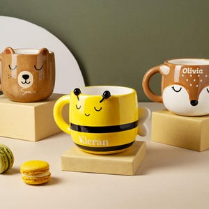 Könnte beinhalten: Drei Keramikbecher mit Tiergesichtern und personalisierten Namen. Die Becher sind gelb, braun und weiß. Die Becher stehen auf einer hellbraunen Oberfläche.