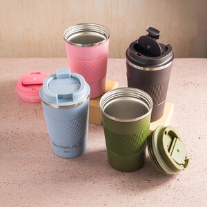 Op de afbeelding: Een assortiment ge&iuml;soleerde reismokken in verschillende kleuren, waaronder roze, blauw, groen en bruin. Elke mok heeft een roestvrijstalen binnenkant en een getextureerde siliconen hoes. Sommige hebben deksels, en &eacute;&eacute;n heeft de tekst "Alex Smith, PhD 2025."
