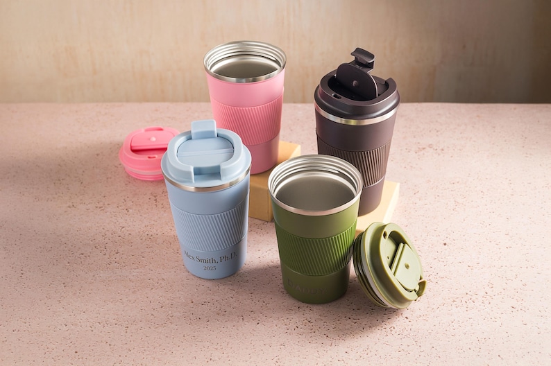 Op de afbeelding: Een assortiment ge&iuml;soleerde reismokken in verschillende kleuren, waaronder roze, blauw, groen en bruin. Elke mok heeft een roestvrijstalen binnenkant en een getextureerde siliconen hoes. Sommige hebben deksels, en &eacute;&eacute;n heeft de tekst "Alex Smith, PhD 2025."