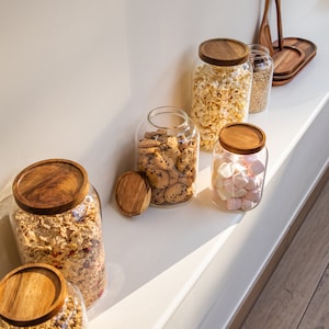 May include: Six glass jars with wooden lids filled with various snacks, including popcorn, cookies, marshmallows, and granola. The jars are arranged on a white shelf with a wooden floor in the background.