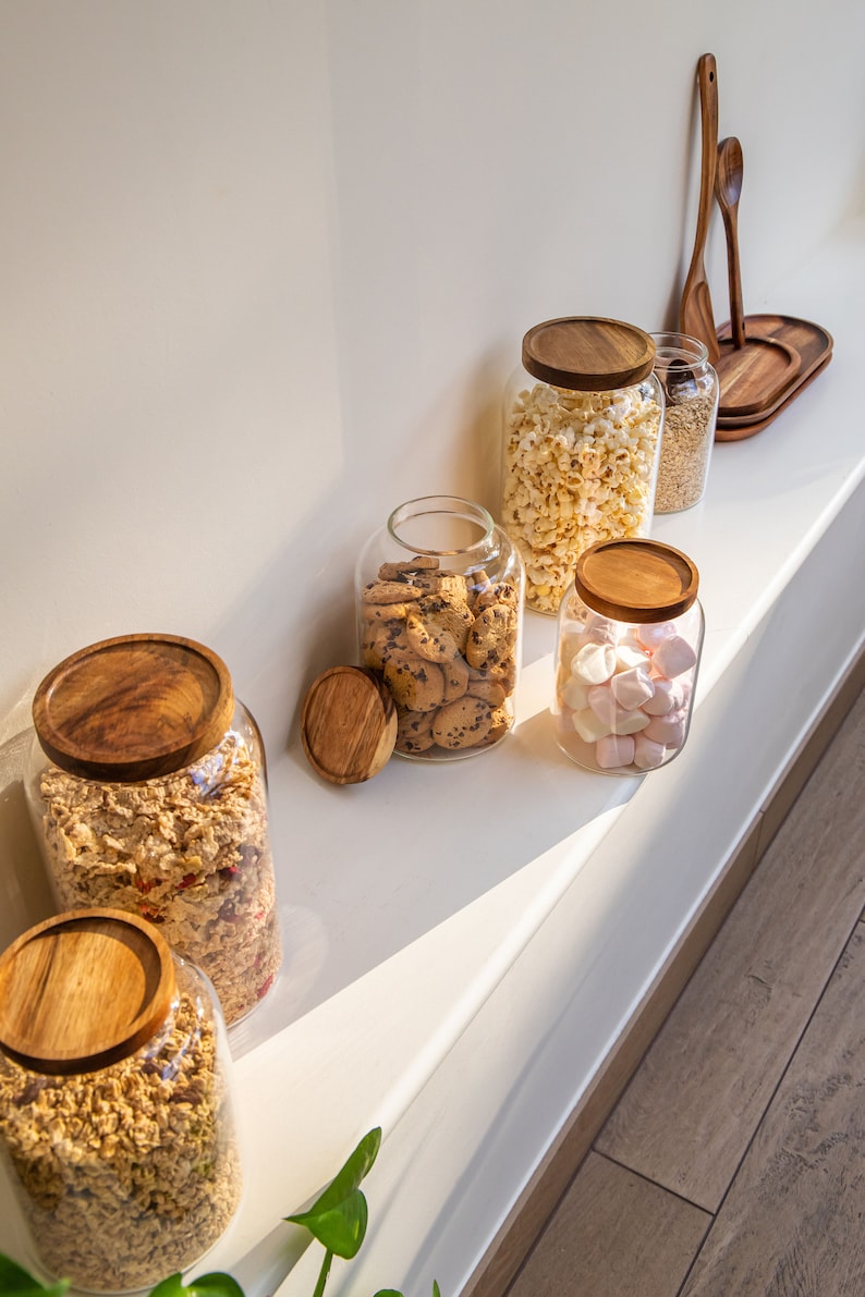 May include: Six glass jars with wooden lids filled with various snacks, including popcorn, cookies, marshmallows, and granola. The jars are arranged on a white shelf with a wooden floor in the background.