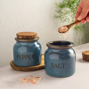 Pu&ograve; includere: Due barattoli blu in ceramica per sale e pepe con coperchi in sughero. Il barattolo a sinistra dice "PEPPER" e quello a destra "SALT". Un cucchiaio di legno contiene sale rosa e un vassoio di legno &egrave; sotto il barattolo del pepe.