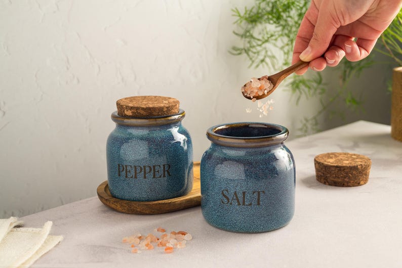 Pu&ograve; includere: Due barattoli blu in ceramica per sale e pepe con coperchi in sughero. Il barattolo a sinistra dice "PEPPER" e quello a destra "SALT". Un cucchiaio di legno contiene sale rosa e un vassoio di legno &egrave; sotto il barattolo del pepe.