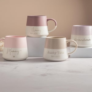 May include: Four ceramic mugs with two-tone designs. One is pink and white with 'Mummy' and a heart, one is mauve and white with 'Nanny EST.2020', one is beige and white with 'Aunty Tina EST.2020', and one is lavender and white with 'Grandma'.