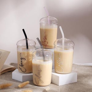 Pu&ograve; includere: Quattro bicchieri in vetro trasparente con coperchi a cupola e cannucce abbinate, riempiti con un liquido beige. I bicchieri presentano etichette con testo in corsivo che riportano Ebony, Silver, Coffee e Rose. Ogni cannuccia &egrave; di un colore diverso: nero, grigio, rosa e bianco.