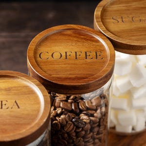 以下が含まれることがあります： 木製の蓋が付いたガラス製の容器が3つあります。蓋には「TEA」、「COFFEE」、「SUGAR」とラベルが貼られています。容器には、お茶、コーヒー豆、砂糖が入っています。