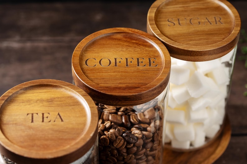 以下が含まれることがあります： 木製の蓋が付いたガラス製の容器が3つあります。蓋には「TEA」、「COFFEE」、「SUGAR」とラベルが貼られています。容器には、お茶、コーヒー豆、砂糖が入っています。