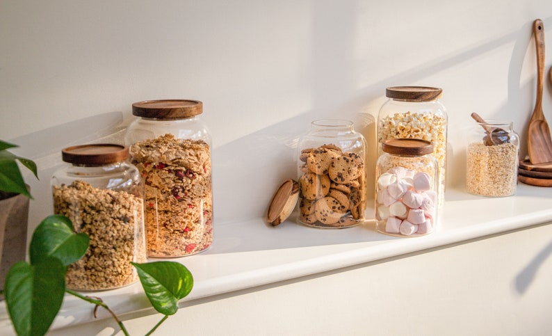 May include: Six glass jars with wooden lids filled with various snacks, including granola, cookies, marshmallows, and popcorn. The jars are arranged on a white shelf.