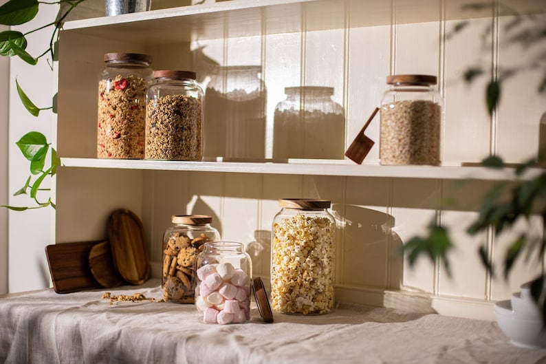 May include: A white kitchen shelf with six glass jars with wooden lids filled with various dry goods, including granola, popcorn, and marshmallows. The jars are on a white shelf with a light wood trim.