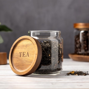 以下が含まれることがあります： 「TEA」と刻まれた木製の蓋が付いたガラス瓶に、茶葉が詰められています。瓶は白い木製の表面に置かれ、近くに茶葉が少し散らばっています。