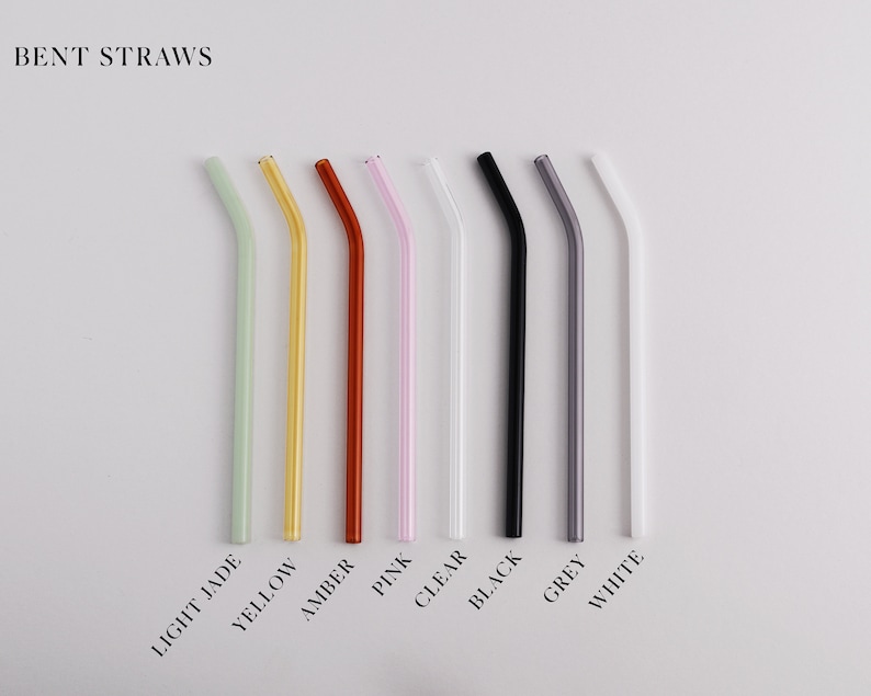 Pu&ograve; includere: Una collezione di otto cannucce in vetro piegate disposte in fila, etichettate BENT STRAWS. I colori includono verde giada chiaro, giallo, ambra, rosa, trasparente, nero, grigio e bianco. Ogni cannuccia presenta una piegatura angolata.