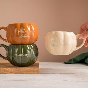 May include: Three pumpkin-shaped ceramic mugs with handles. The top mug is orange with the text Hello Autumn. The bottom mug is dark green with the text Mummy. The third mug is cream-coloured with the text Pumpkin Spice Latte in white script.