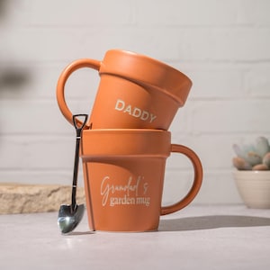 Pode incluir: Duas canecas cor de terracota empilhadas, parecendo vasos de flores, com alças. Uma caneca diz "DADDY", a outra "Grandad's garden mug". Uma pequena colher em forma de pá está ao lado. Suculentas em vasos brancos e bege ao fundo.