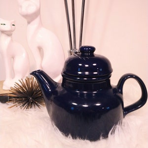 May include: A navy blue ceramic teapot with a matching lid sits on a fluffy white surface. Two white cat figurines and a gold starburst decoration are in the background. Tall silver rods are also visible.