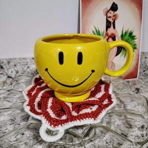Puede incluir: Una taza amarilla con una cara sonriente negra, colocada sobre un posavasos de ganchillo rojo y blanco. Una ilustración de estilo vintage de una mujer en un entorno tropical está en el fondo.