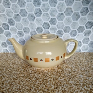 Może przedstawiać: Kremowy ceramiczny czajnik z pasującą pokrywką i uchwytem. Czajnik posiada serię pomarańczowych i zielonych kwadratowych i diamentowych wzorów. To artykuł kuchenny.