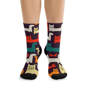 Op de afbeelding: Een paar zwarte sokken met een kleurrijk patroon van katten en dinosaurussen. De katten hebben gesloten ogen en zijn in verschillende kleuren, waaronder rood, geel, groen en blauw. De dinosaurussen zijn in tinten groen, blauw en geel.