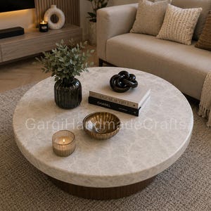 May include: A round coffee table with a light-colored stone top and a dark wood base. On the table are a dark glass vase with greenery, a stack of two books titled Live Beautiful and Chanel, a black knot sculpture, a gold bowl, and a candle.