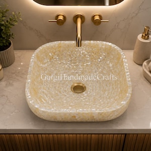 May include: A square vessel sink with a textured, iridescent mother-of-pearl finish in shades of cream and pale yellow. The sink features a central circular drain cover in a brushed gold finish. The text GargiHandmadeCrafts is visible.