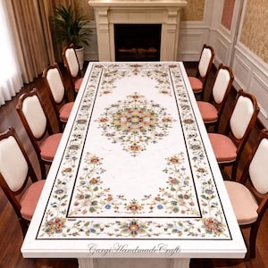 Op de afbeelding: Een grote witte marmeren eettafel met ingewikkelde bloemeninleg in verschillende kleuren. De tafel is omringd door houten stoelen met witte rugleuningen en roze zittingen. De tafel is ongeveer 305 cm lang.
