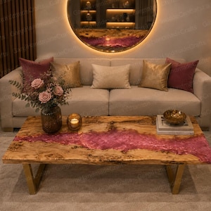 May include: A rectangular coffee table featuring a natural live-edge wood slab with a central river inlay of translucent pink resin. The table is supported by two angular gold-toned metal legs and is positioned in front of a beige sofa with cushions.