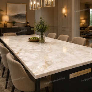 Puede incluir: Una mesa de comedor rectangular con una superficie de piedra blanca pulida con vetas doradas, sostenida por una base oscura. Una placa rectangular con el texto GargiHandmadeCrafts está fijada en el lateral. Sillas de color beige rodean la mesa.