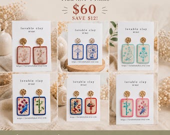May include: A collection of six pairs of rectangular mahjong tile earrings displayed on white cards labeled Lovable Clay by Kat. Each pair features unique illustrations including floral patterns, birds, bamboo, and lanterns with gold-toned hardware.