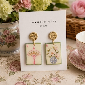 May include: A pair of rectangular clay earrings attached to a white display card labelled "lovable clay BY KAT". Each earring features a textured gold-tone stud and a cream-coloured rectangular pendant with a green border, displaying floral art.
