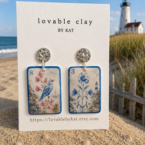 K&ouml;nnte beinhalten: Ein Paar rechteckige H&auml;ngeohrringe auf einer wei&szlig;en Pr&auml;sentationskarte mit der Aufschrift lovable clay BY KAT. Jeder Ohrring hat einen blauen Rand und eine Vintage-Illustration. Einer zeigt einen blauen Vogel, der andere eine blaue Blume.