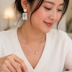 May include: Gold-toned earrings with a textured circular top and a rectangular pendant featuring a bamboo design. A delicate gold necklace with a flower pendant is also visible. The image showcases a Mahjong tile with a bamboo illustration.
