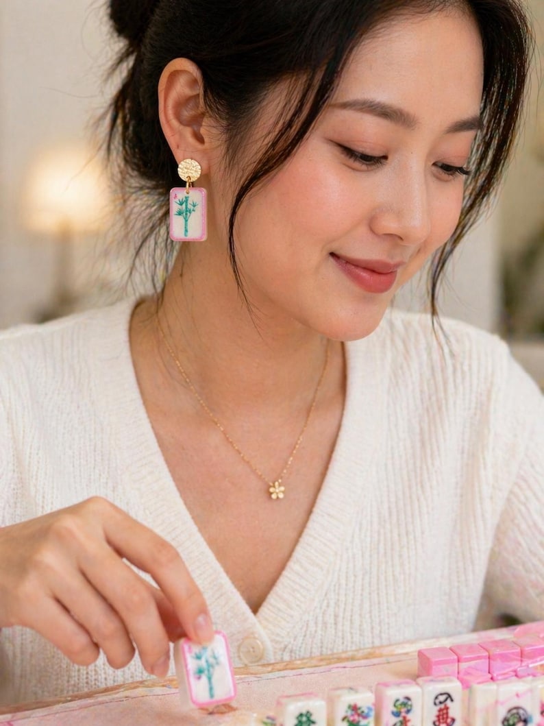 May include: Gold-toned earrings with a textured circular top and a rectangular pendant featuring a bamboo design. A delicate gold necklace with a flower pendant is also visible. The image showcases a Mahjong tile with a bamboo illustration.