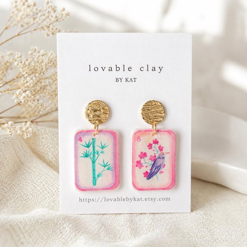 May include: A pair of handmade earrings featuring a gold disc stud and a rectangular pendant with a pink border. One pendant has a bamboo design, the other a bird and floral design. The text "lovable clay" and the Etsy shop URL are visible.