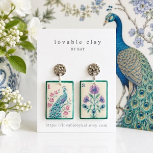Pode incluir: Um par de brincos retangulares pendentes num cartão branco com a etiqueta lovable clay BY KAT. O brinco esquerdo apresenta um pavão e o número 1, enquanto o direito mostra uma flor roxa e o número 3, ambos com bordas verde-azuladas.