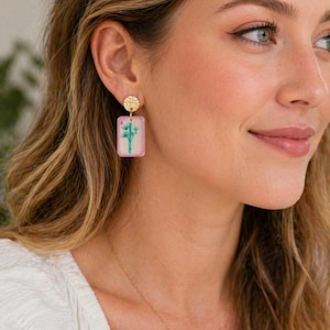 May include: Gold-colored stud earrings with a textured circular top and a rectangular, pink-edged pendant. The pendant features a bamboo design in green and blue. A delicate gold necklace is also visible.