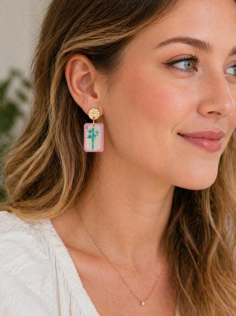May include: Gold-colored stud earrings with a textured circular top and a rectangular, pink-edged pendant. The pendant features a bamboo design in green and blue. A delicate gold necklace is also visible.