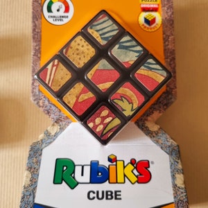 May include: A Rubik's Cube with a patterned design. The cube is black with a colourful pattern on each side. The packaging is orange and black with the words "Rubik's Cube" and "World's No.1 Puzzle" printed on it.