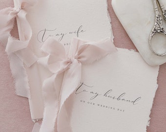 May include: Two white paper journals with a pink ribbon tied around each. The journal on the left has the text "To my wife" written on it. The journal on the right has the text "To my husband on our wedding day" written on it.