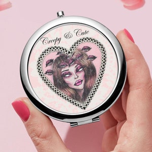 May include: A silver compact mirror with a heart-shaped design. The heart features a pink and white background with a black and white illustration of a woman with eyes surrounded by lips. The text "Creepy & Cute" is printed above the heart.