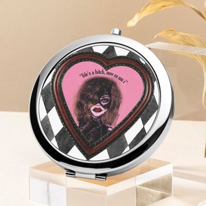 May include: A silver compact mirror with a black and white chequered design. The mirror has a heart-shaped opening with a pink background and a woman wearing a mask and a black outfit. The text "Life's a bitch, now so am I" is printed on the heart.