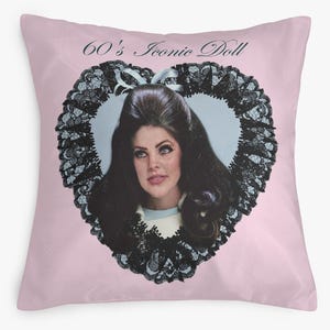 May include: A pink cushion with a portrait of a woman with dark hair, framed by a black lace heart. The text "60's Iconic Doll" is arched above the heart. The cushion has a solid pink background.