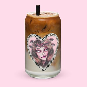 Puede incluir: Una lata de vidrio con un diseño de corazón rosa que presenta a una mujer con múltiples ojos y el texto "creepy & cute". La lata está llena de café helado.