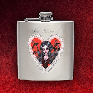 Puede incluir: Una petaca de plata con una ilustración en blanco y negro de una mujer con el pelo largo y negro y un corazón rojo con ribete de encaje. El texto "Death Imitates Art" está impreso en la petaca.